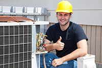 HVAC technician kneeling by a condenser wearing a yellow hard hat smiling and giving a thumbs up